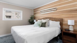 Ein Schlafzimmer mit einem großen Bett, einem Nachttisch mit Lampe, einem Surfbrett als Wanddekoration und einem Fenster mit Jalousien.