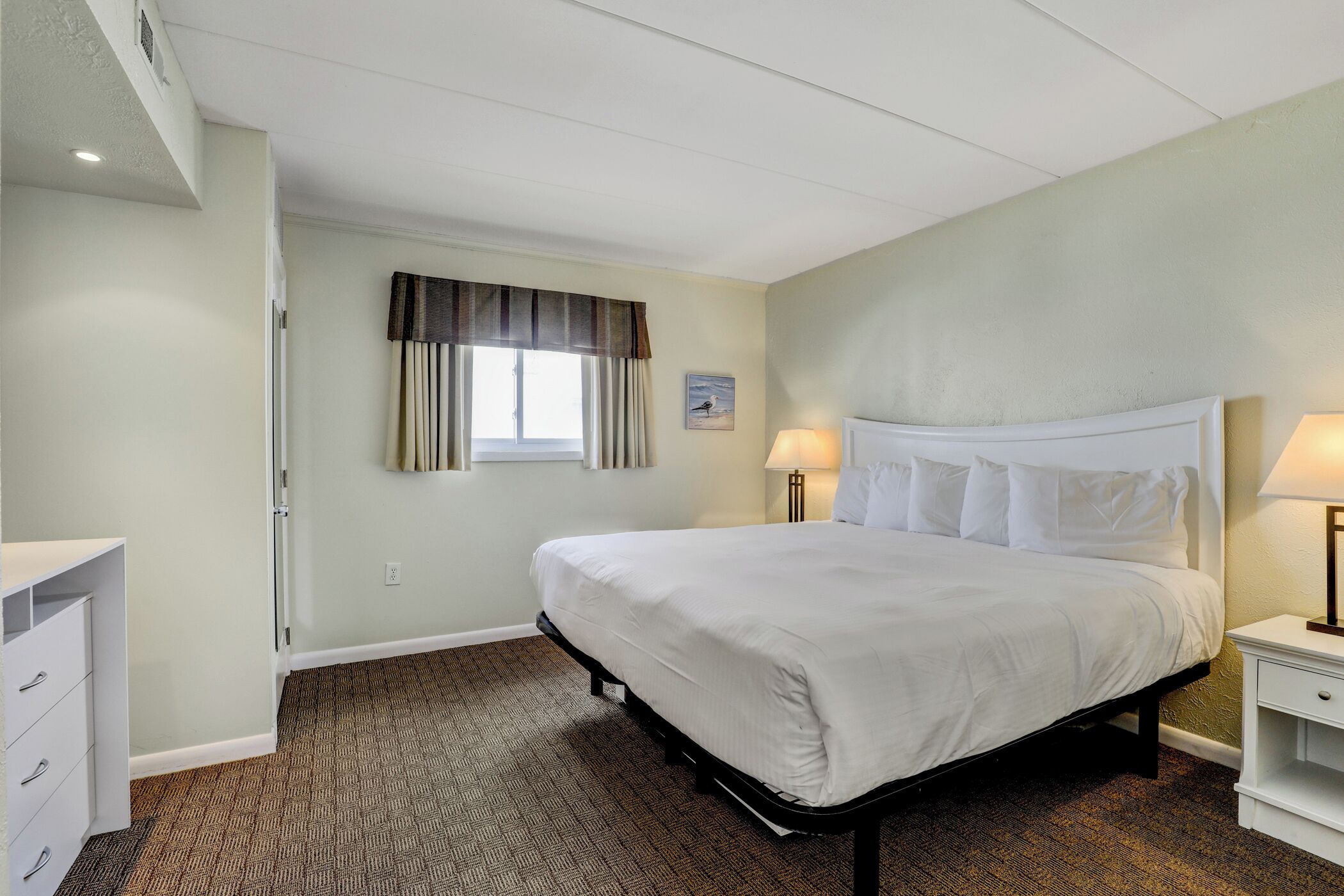 A hotel room with a large bed, two bedside lamps, a window with curtains, and a small bedside table.