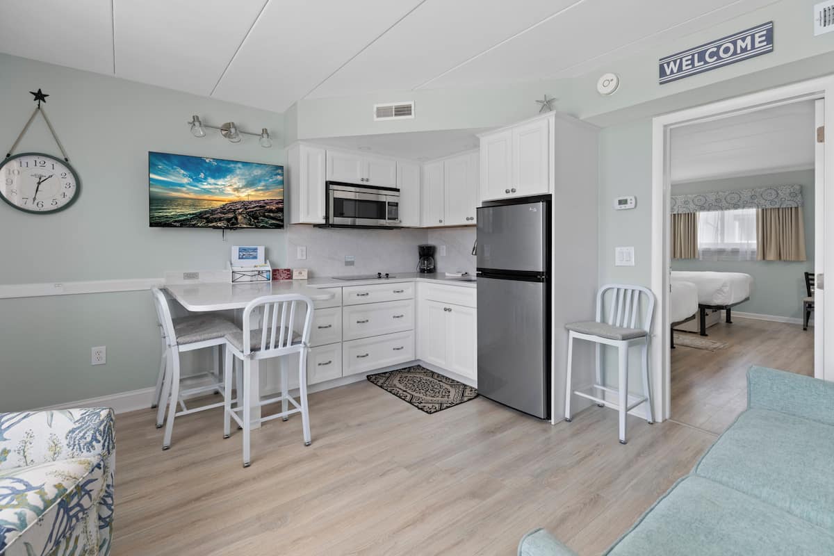 A compact kitchen with white cabinets, a stainless steel refrigerator, a microwave, and a dining area with two white chairs.