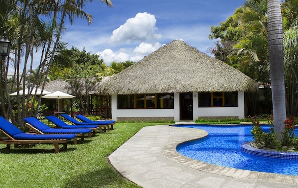 A thatched-roof building with a pool and lounge chairs.