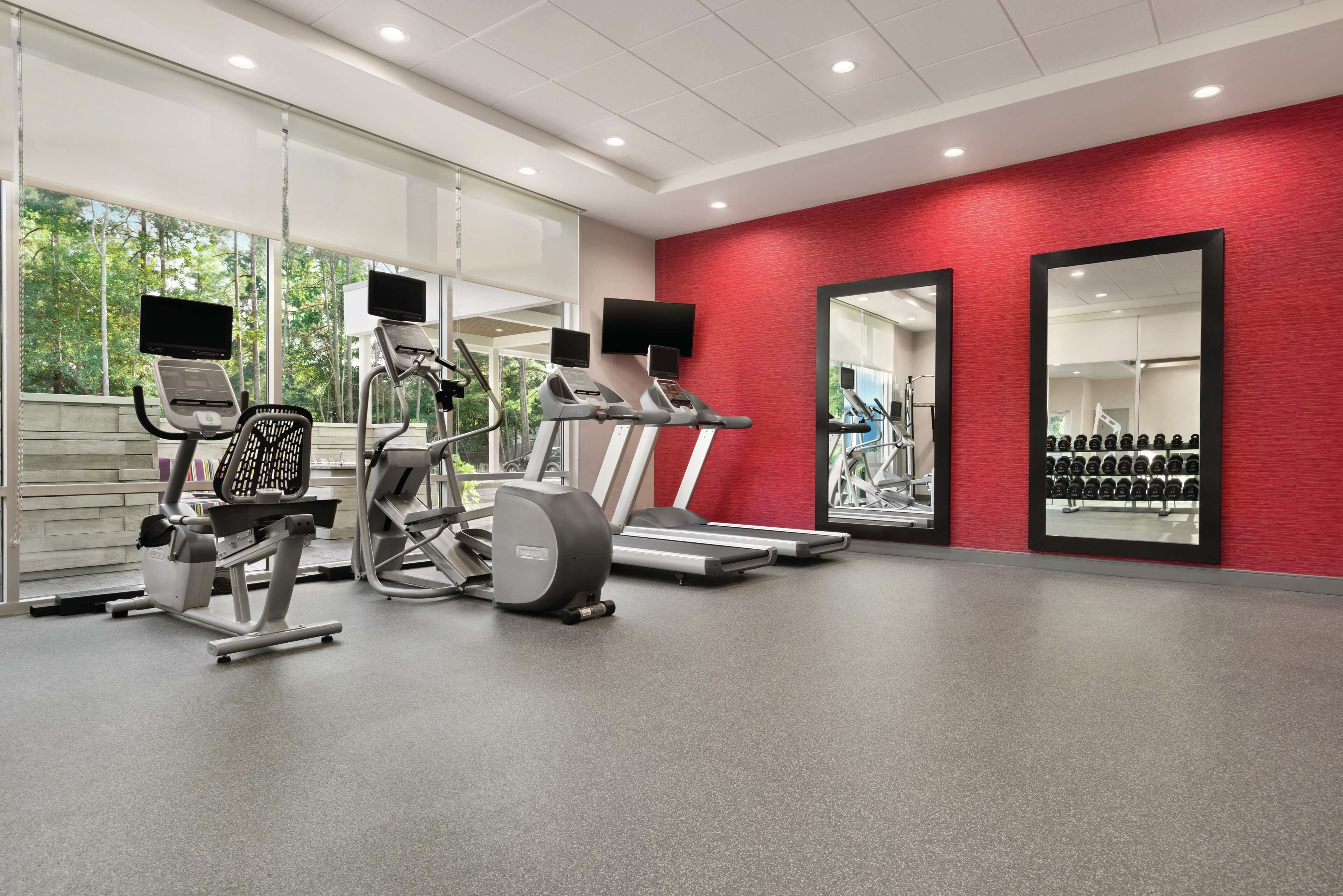 Une salle de sport équipée de machines de cardio, avec un mur rouge et un grand miroir.
