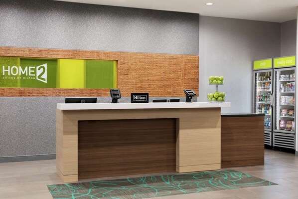 A reception desk with a modern design, a green 'HOME2' sign, a phone, and a 'Hilton Honors' plaque. There are two refrigerators labeled 'cold' and 'really cold' in the background. - Home2 Suites by Hilton Columbia Harbison (Irmo)