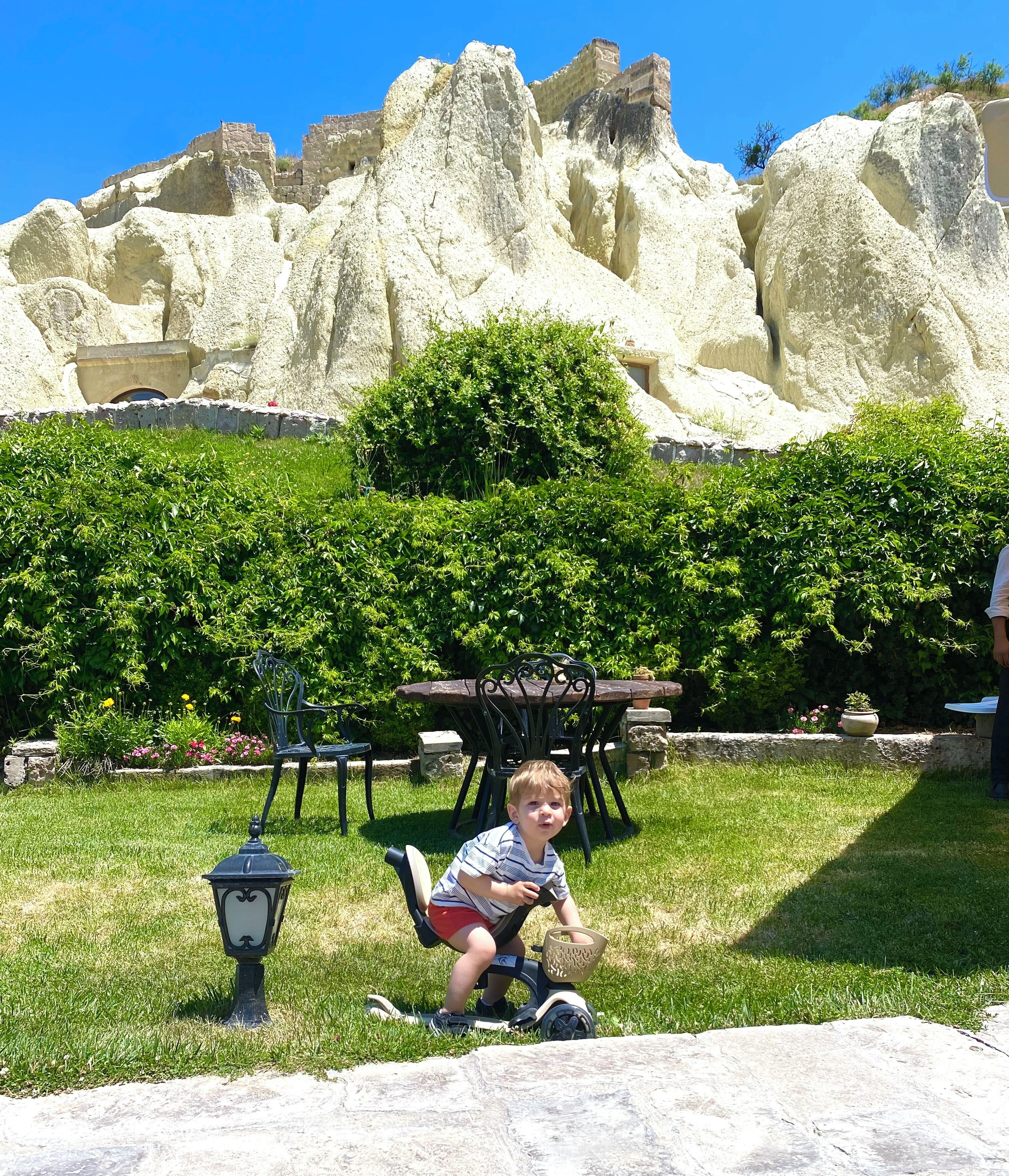 A child on a tricycle in a grassy area with a stone wall and a table with chairs in the background.