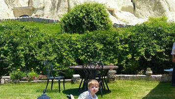A child on a tricycle in a grassy area with a stone wall and a table with chairs in the background.