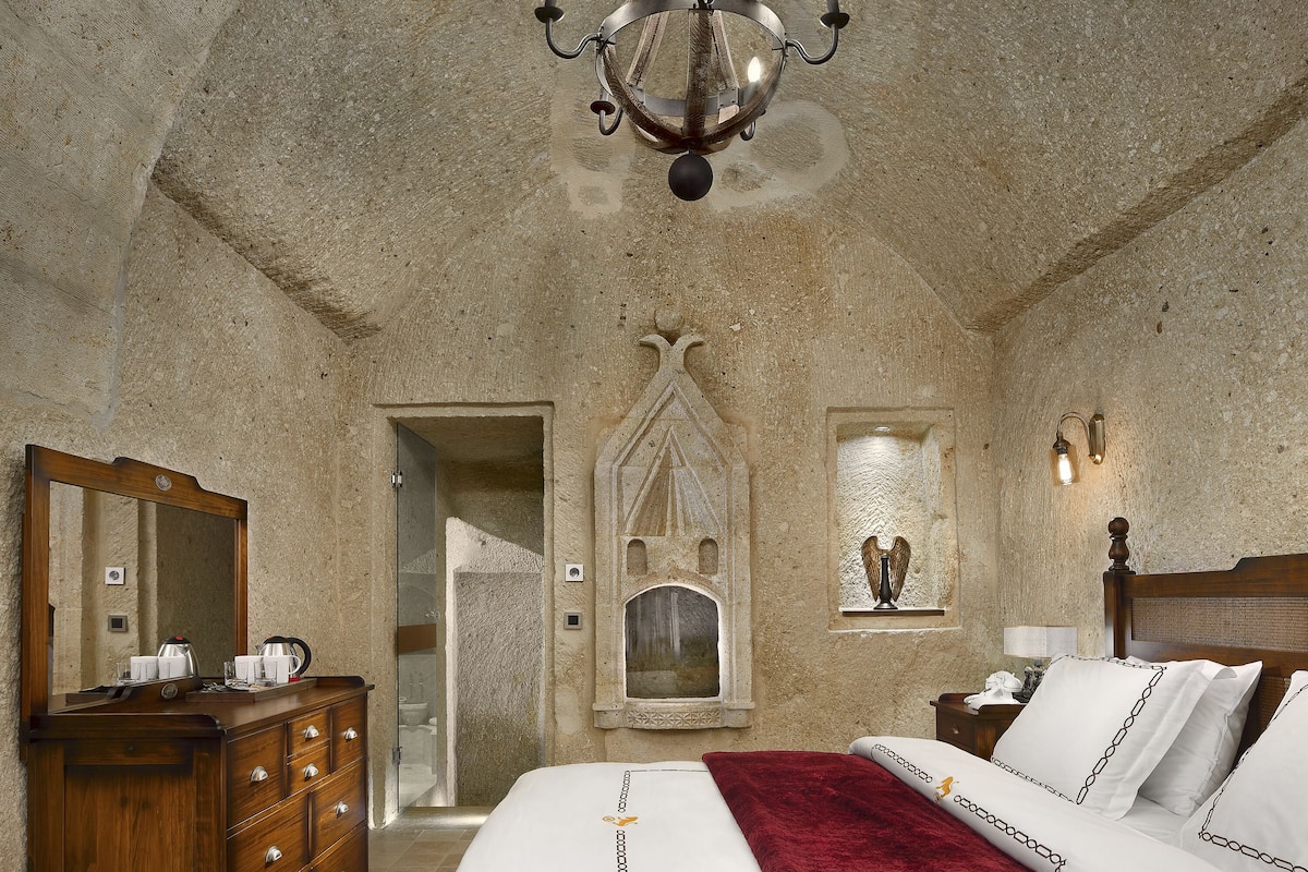 A bedroom with a large bed, a wooden dresser, a chandelier, and a stone wall.