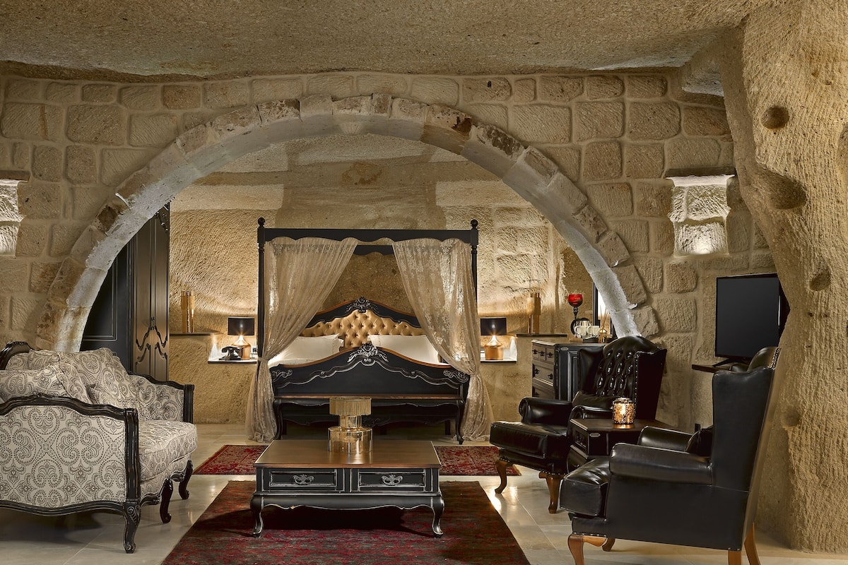 A bedroom with a four-poster bed, a sitting area with a sofa and armchair, a television, and a stone wall arch.