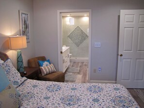 A bedroom with a bed, a chair, a lamp, a door, and a bathroom visible through an open door. - Windancer by Crystal Waters (Destin)
