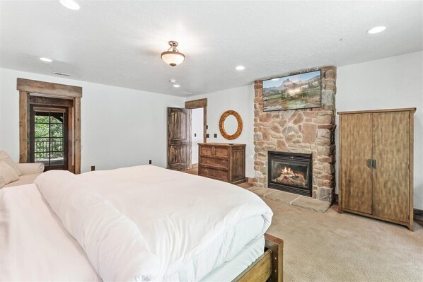 A bedroom with a large bed, a stone fireplace, wooden furniture, and a door leading to another room. - HIDDEN MOUNTAIN LUXURY LODGE, CLOSE TO SKI RUNS, HOT TUB, THEATER, SALOON (Sundance)