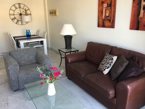 A living room with a brown leather sofa, a grey armchair, a glass coffee table with a vase of flowers, a wall clock, and two lamps.