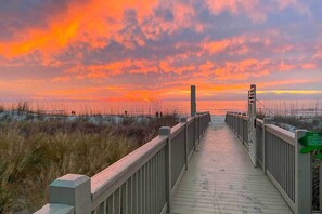 En træpromenade der fører til en strand med solnedgang i baggrunden.