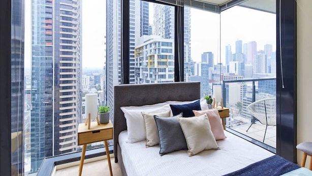 A bedroom with a large bed, a bedside table with a lamp, and a view of a cityscape through large windows. - Penthouse Apartment in Melb CBD Perfect Location (Melbourne)