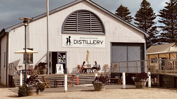 A distillery building with a large arched window, outdoor seating area, and potted plants.