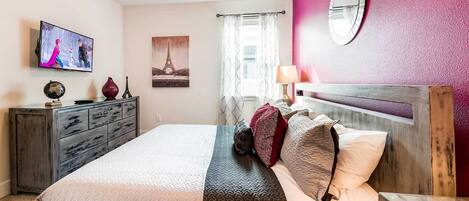 A bedroom with a bed, a dresser, a television, a painting of the Eiffel Tower, and a mirror on the wall.