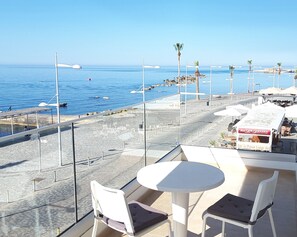 A terrace with a table and chairs overlooking a beach with palm trees and a clear blue sky.