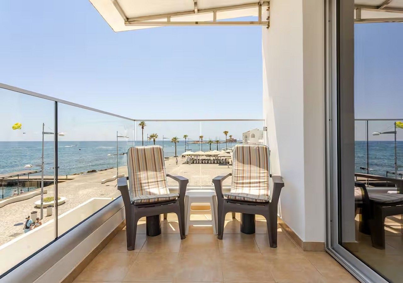 A balcony with a view of the beach, two chairs, and a table.