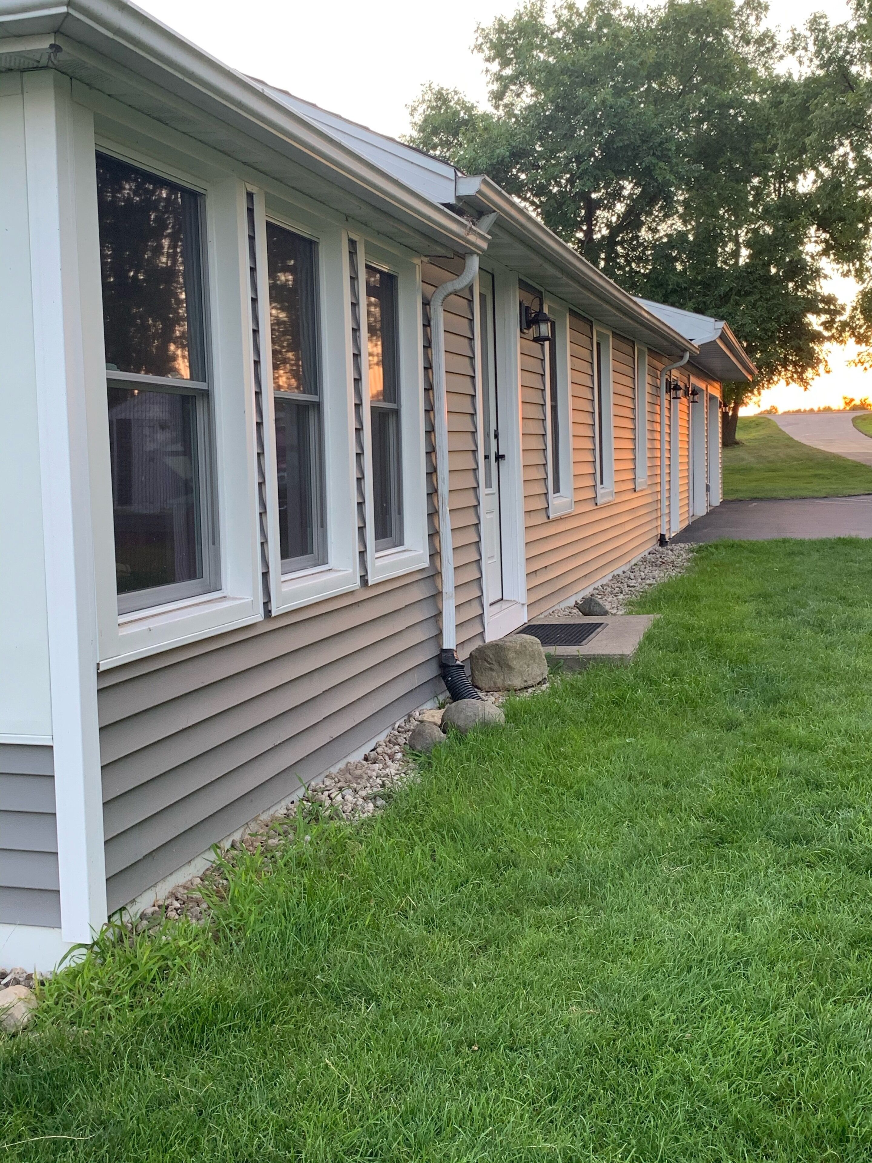 A single-story house with a grey siding exterior, multiple windows with white trim, and a small front yard with grass and rocks.
