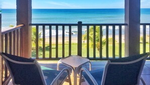 Ein Balkon mit zwei Stühlen und einem kleinen Tisch, der auf einen Strand mit klarem, blauem Wasser und eine Rasenfläche mit Palmen blickt.