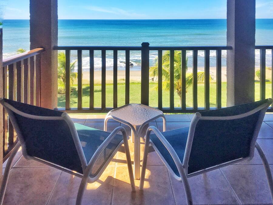 A balcony with two chairs and a small table, overlooking a beach with clear blue water and a grassy area with palm trees.