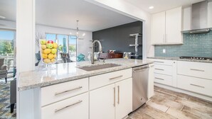 A modern kitchen with white cabinets, a stainless steel dishwasher, and a blue-tiled backsplash. - Remodeled Harbor Condo Steps to Lake MI (Manistee)