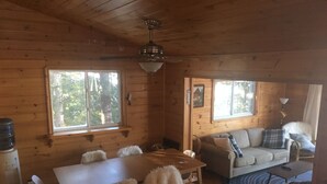 Interior de uma cabana de madeira com uma mesa de jantar, cadeiras, um sofá e um ventilador de teto.