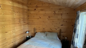 Quarto de cabana de madeira com cama, mesa de cabeceira, luminária e cortinas.