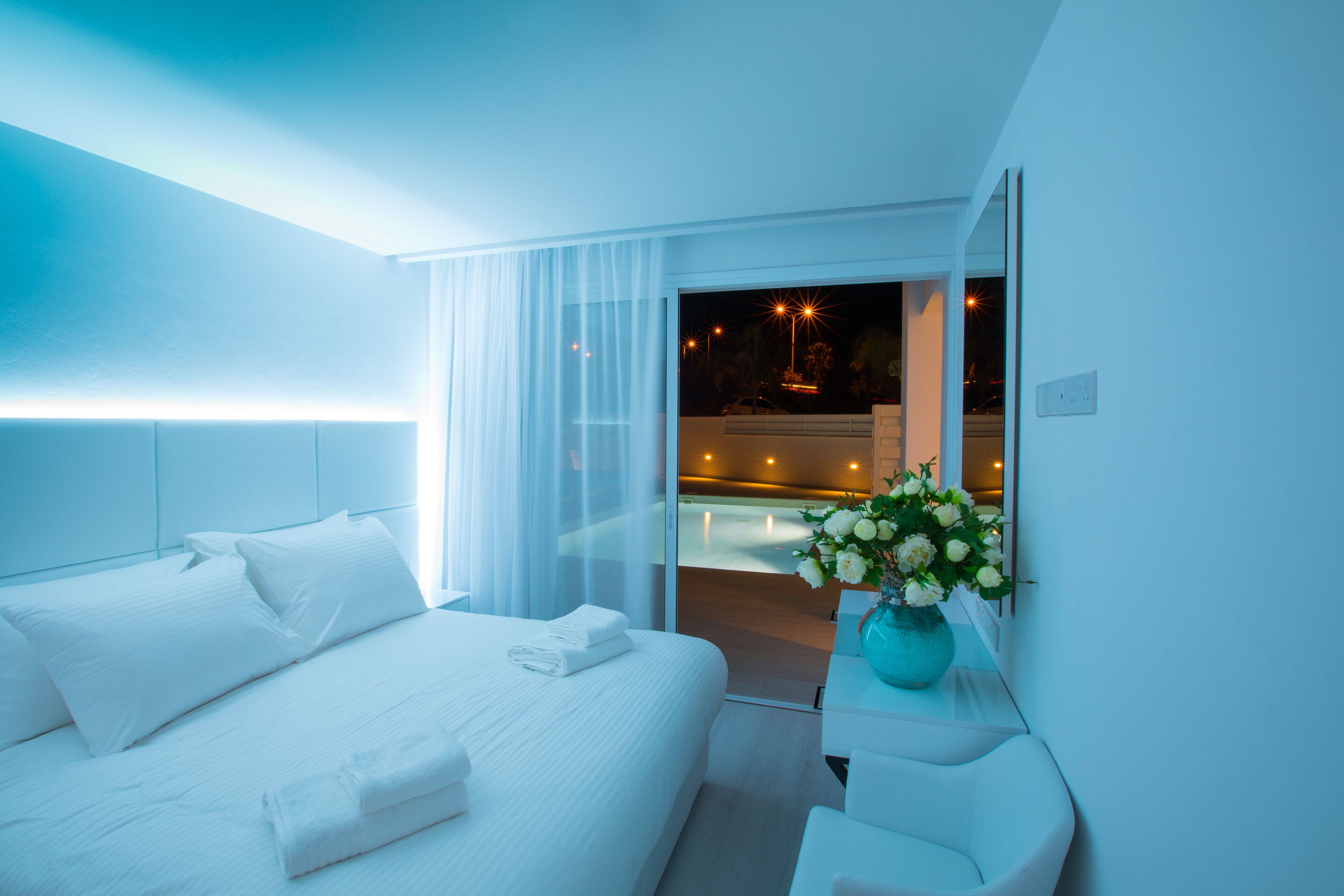 A modern hotel room with a large bed, a bedside table with a vase of flowers, and a view of a pool area at night.