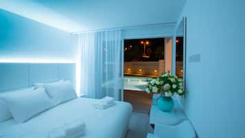 A modern hotel room with a large bed, a bedside table with a vase of flowers, and a view of a pool area at night.