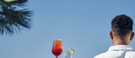 A person holding a tray with a drink and a glass of water, with a clear blue sky and a palm tree in the background.