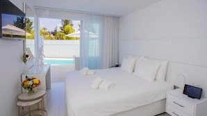 A modern hotel room with a large bed, a bedside table with a digital clock, a small round table with a vase of flowers, and a view of a pool and palm trees through the window. - Hotel Napa Suites - Adults Only (Ayia Napa)