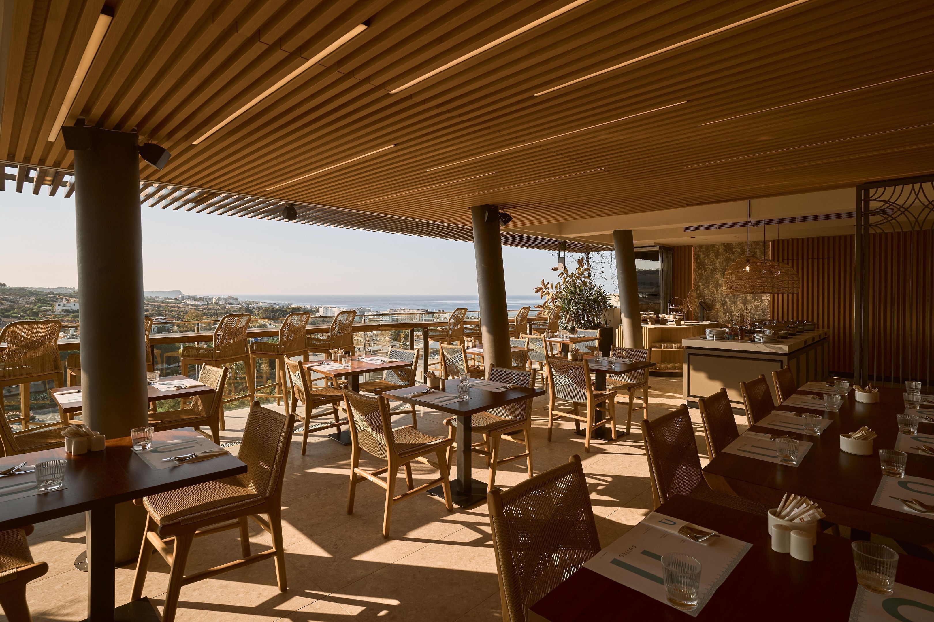 An outdoor dining area with wooden tables and chairs, a view of the city, and a modern ceiling design.