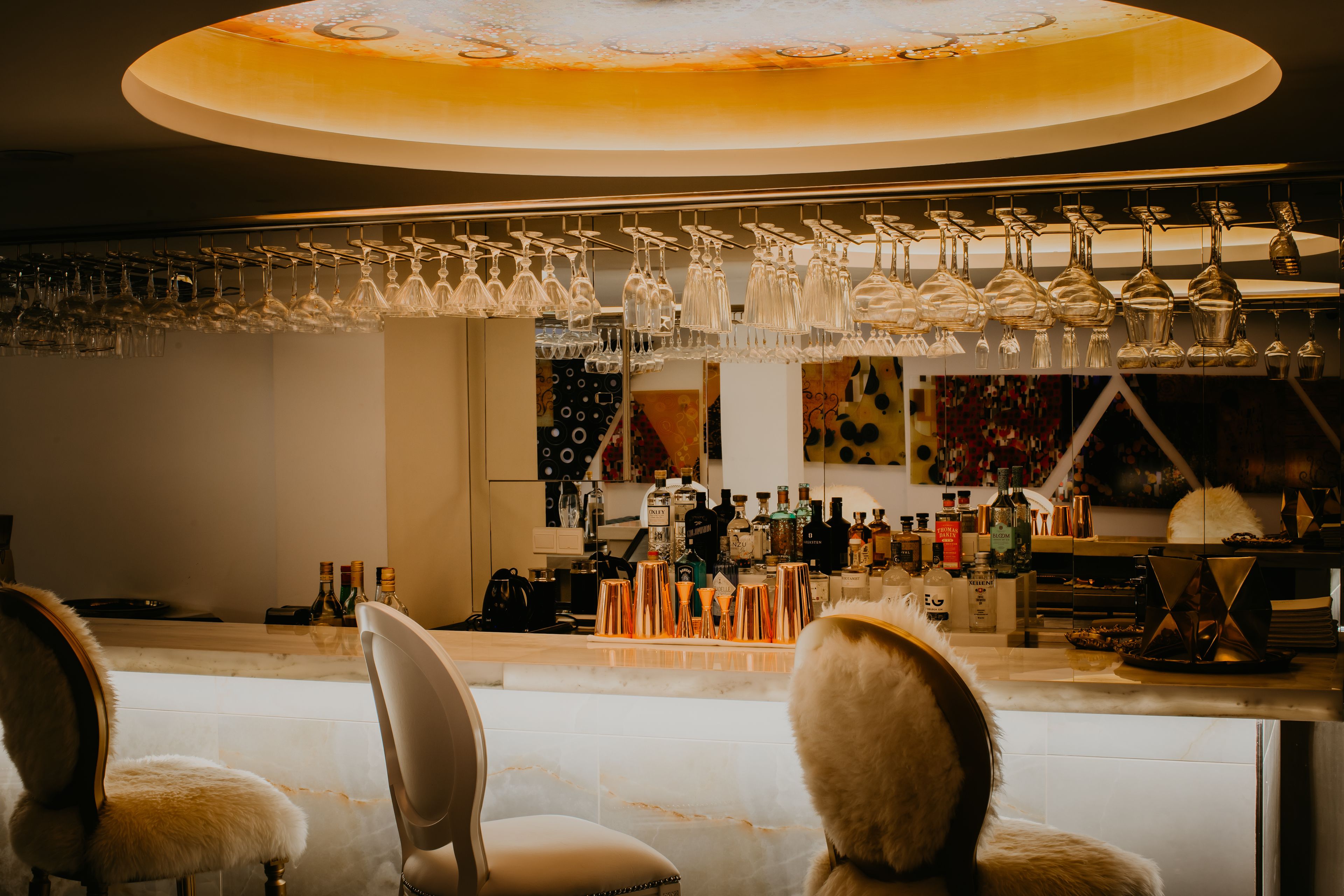 Un espace bar agrémenté de plusieurs verres à vin suspendus, d’un comptoir où sont exposées différentes bouteilles, et de deux fauteuils confortables.