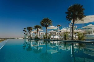 Un área de piscina con sillones de descanso y sombrillas, rodeada de palmeras y un cielo azul despejado.