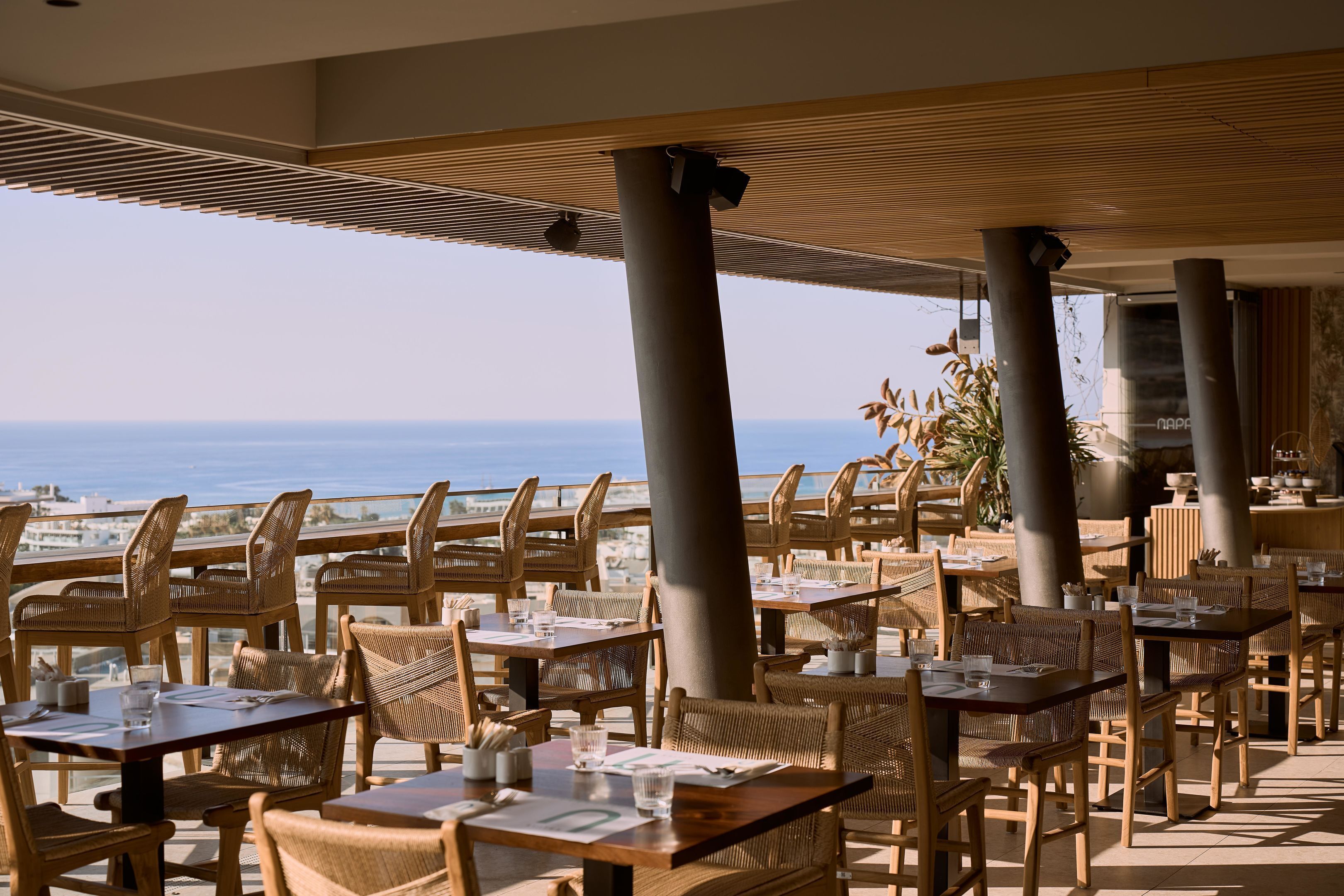 A restaurant with outdoor seating, wooden tables and chairs, and a view of the ocean.