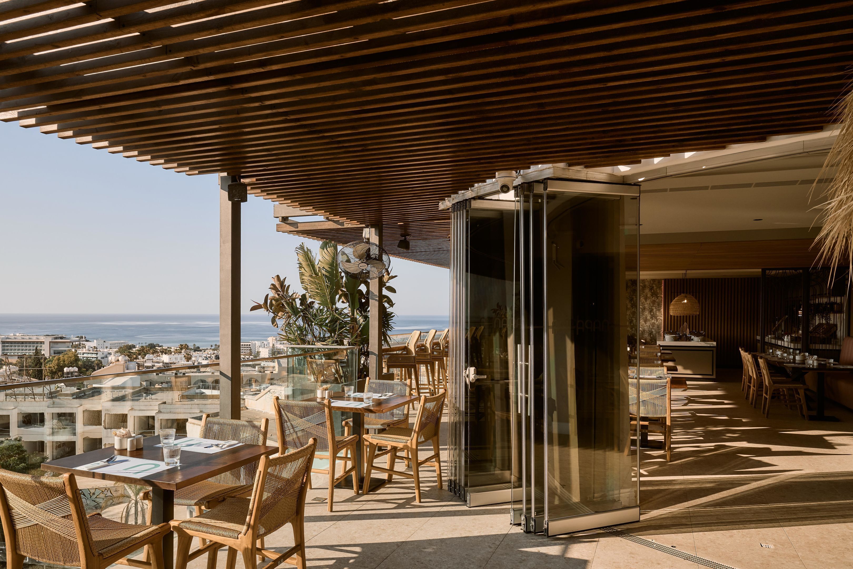 Una terraza con mesas y sillas, un ventanal de vidrio y vista al mar.
