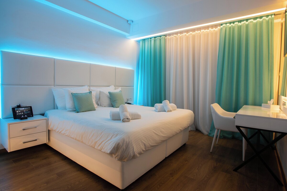 Une chambre d’hôtel moderne avec un lit bien fait, une table de chevet avec une horloge, un bureau avec une chaise et une grande fenêtre avec des rideaux.