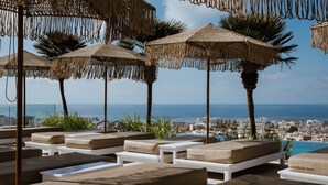 A rooftop lounge area with multiple sunbeds and thatched umbrellas, overlooking a city and the sea. - Hotel Napa Suites - Adults Only (Ayia Napa)