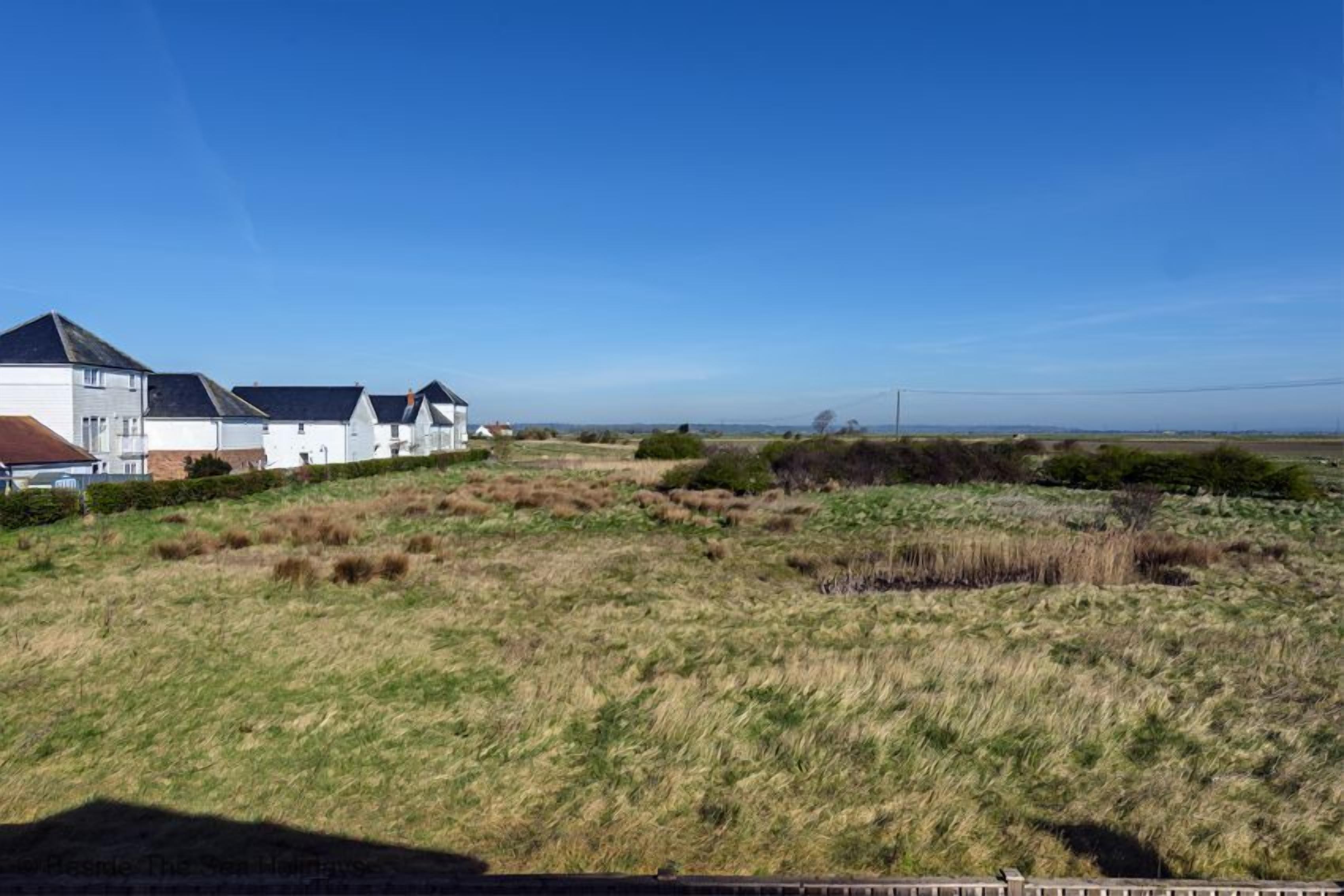 Een landelijk landschap met een rij witte huizen met donkere daken, een grasveld en een heldere blauwe lucht.