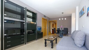 Ein modernes Wohnzimmer mit einem grauen Sofa, einem Couchtisch und einem Fernseher. Es gibt eine Essecke mit Tisch und Stühlen sowie eine Küchenzeile mit Schränken und einem Kühlschrank.