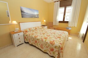 Un dormitorio con cama, mesitas de noche, una silla, un banco, un cuadro y una ventana con cortinas.
