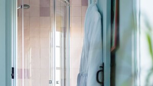 A glass-enclosed shower area with a tiled floor, a wooden chair with a blue cushion, and a white robe hanging on a rack.
