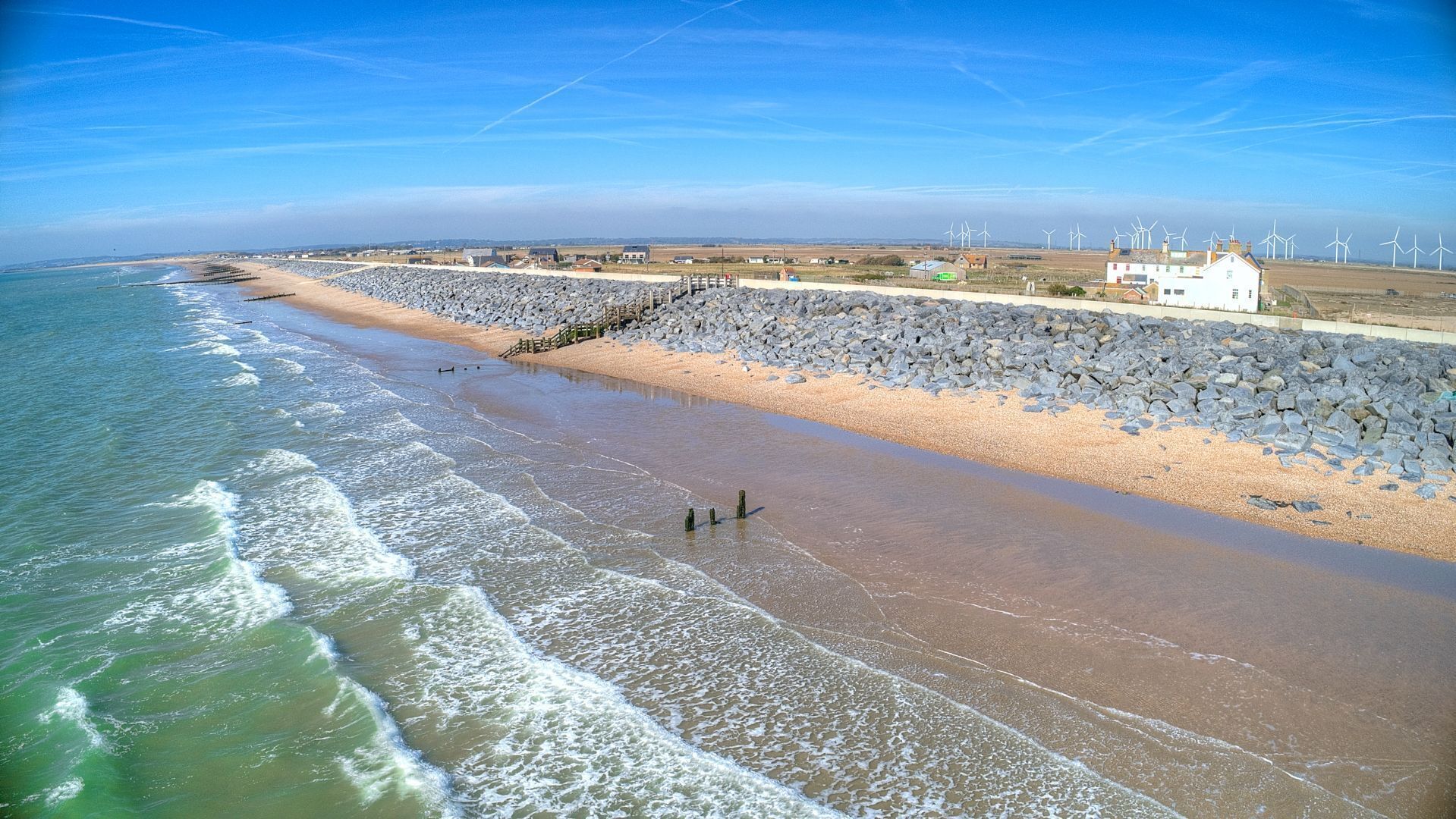 A coastal beach with a rocky breakwater, a sandy shore, and wind turbines in the distance.
