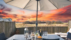 Un espace de restauration en plein air avec une table dressée pour deux personnes, située sous un grand parasol et offrant une vue panoramique sur un magnifique coucher de soleil au-dessus d’un champ.