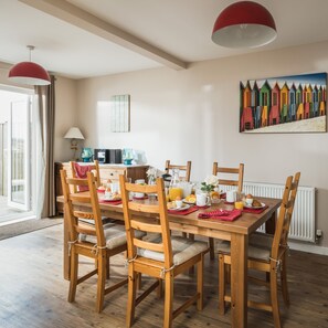 A dining area with a wooden table set for breakfast, surrounded by wooden chairs with cushions. A painting of colorful beach huts hangs on the wall.