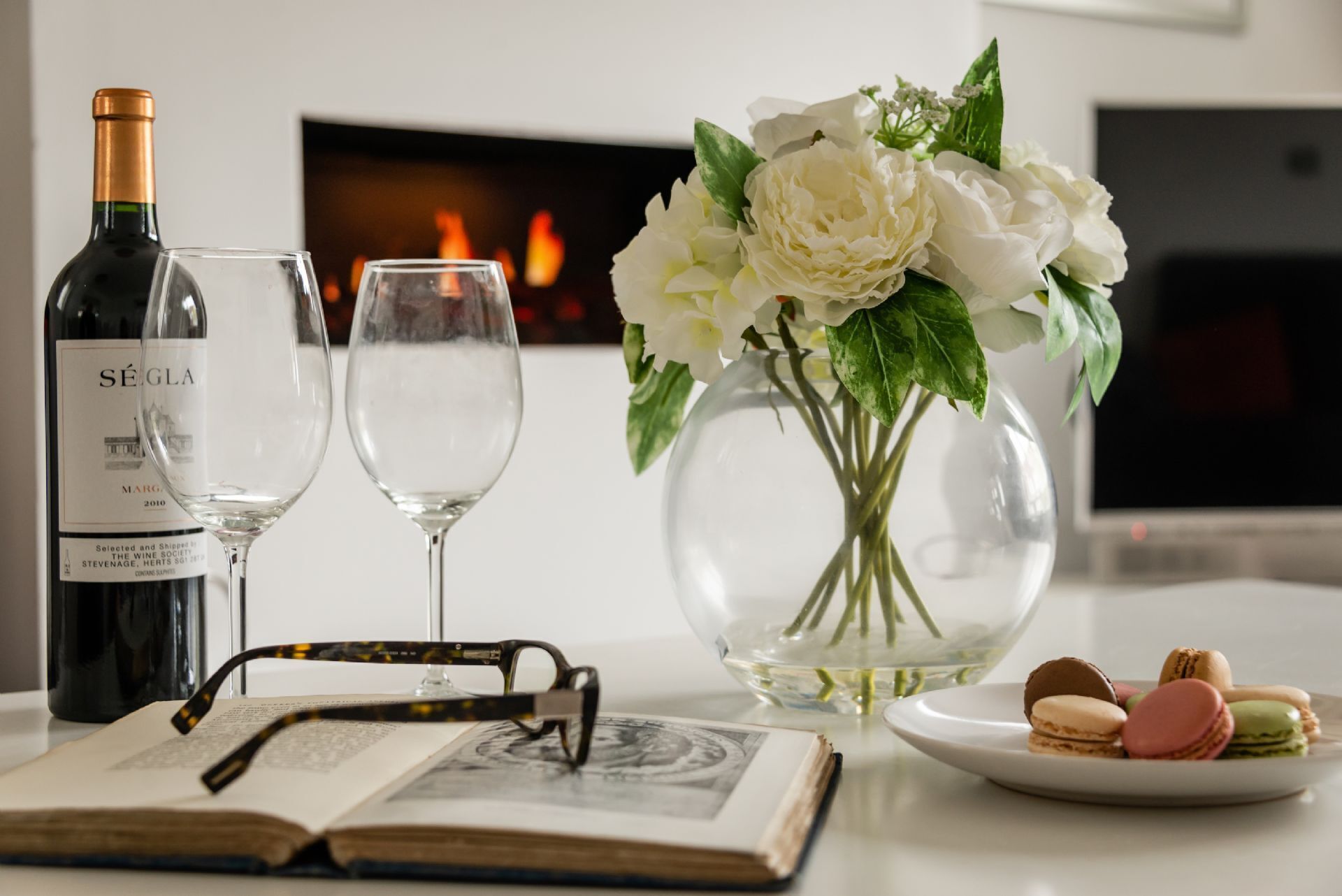 A table with a bottle of wine, two glasses, a book, a pair of glasses, a vase with flowers, and macarons.