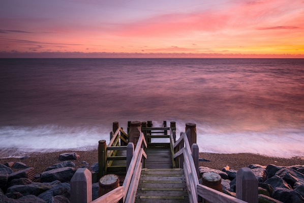 Une jetée en bois menant à une plage rocheuse, avec un coucher de soleil en arrière-plan.