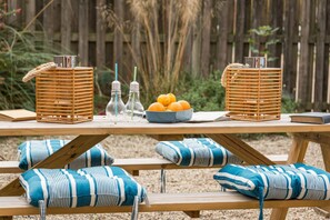 Une table de pique-nique en bois, accompagnée de deux paniers en osier, d’un saladier rempli d’oranges et de deux bouteilles en verre avec des pailles rayées.
