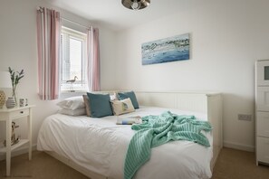 Une chambre à coucher avec un lit blanc, des rideaux à rayures, un tableau représentant un paysage côtier et une table de chevet blanche agrémentée d’une lampe et d’objets décoratifs.
