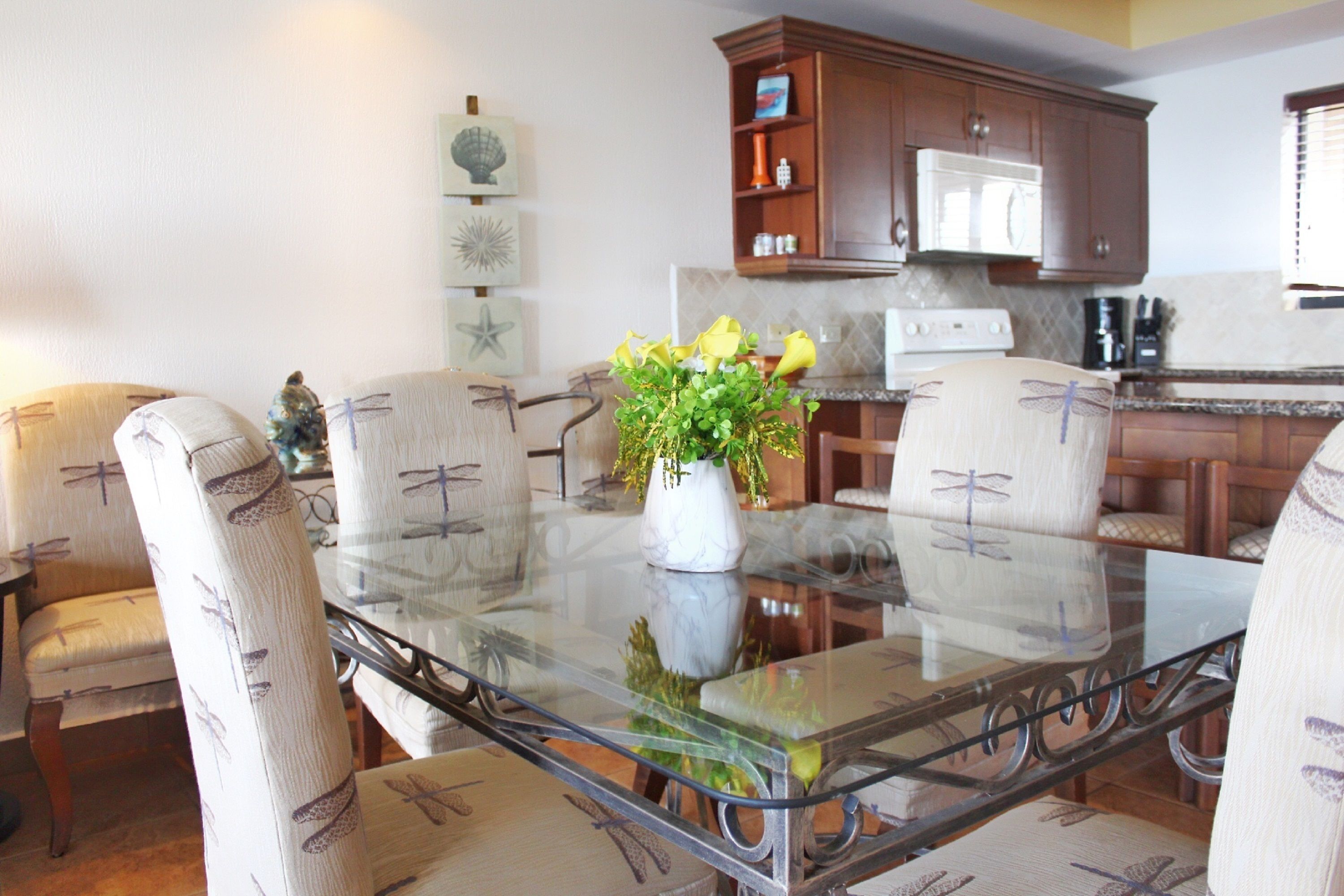 A dining area with a glass table surrounded by chairs, a white vase with flowers, and wooden cabinets in the background.