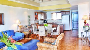 A spacious living area with a blue sofa, wicker furniture, a dining table with chairs, and a kitchen in the background. - Suite San Juan 135 Gran Pacifica Resort (Villa El Carmen)