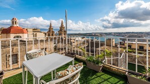 Una terraza en la azotea con una mesa y sillas blancas, plantas en macetas y vistas de edificios históricos y una masa de agua.